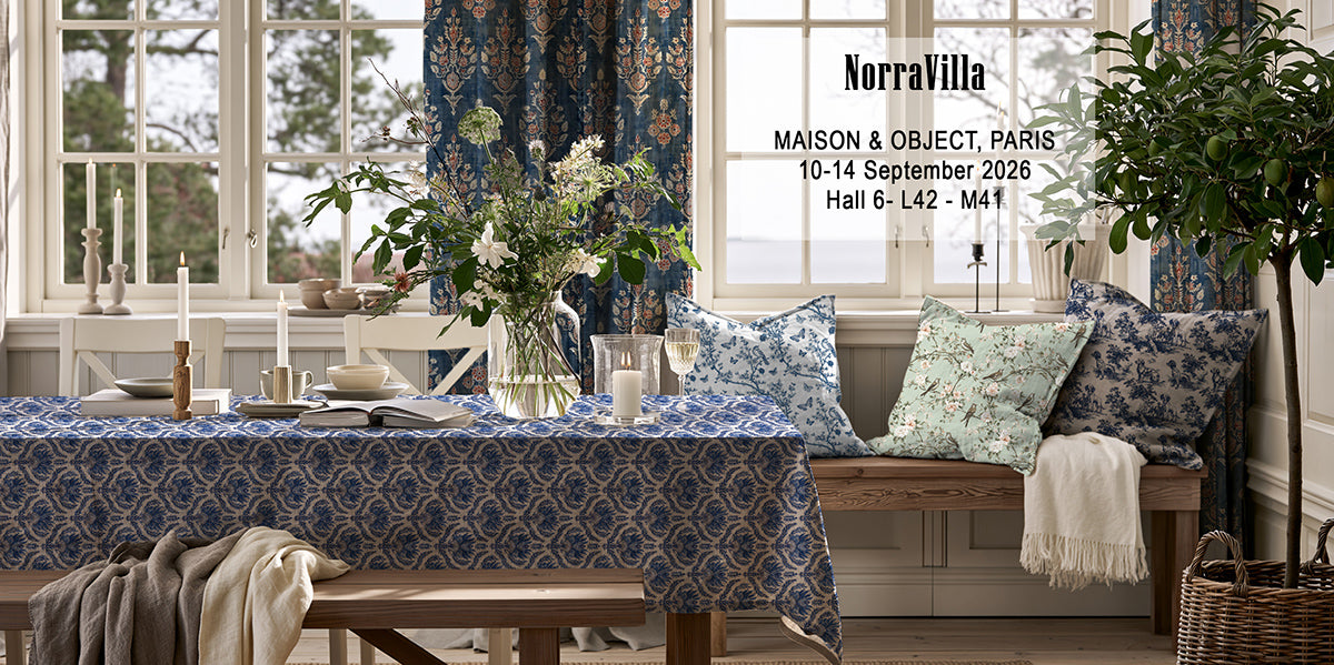 A styled interior scene showing blue patterned tablecloth, pillows, and curtains in a dining or living room setting, with a bench, flowers, candles, and large windows. Text on the right reads 'NorraVilla' with address and event details - Maison & object Paris 2026 September,